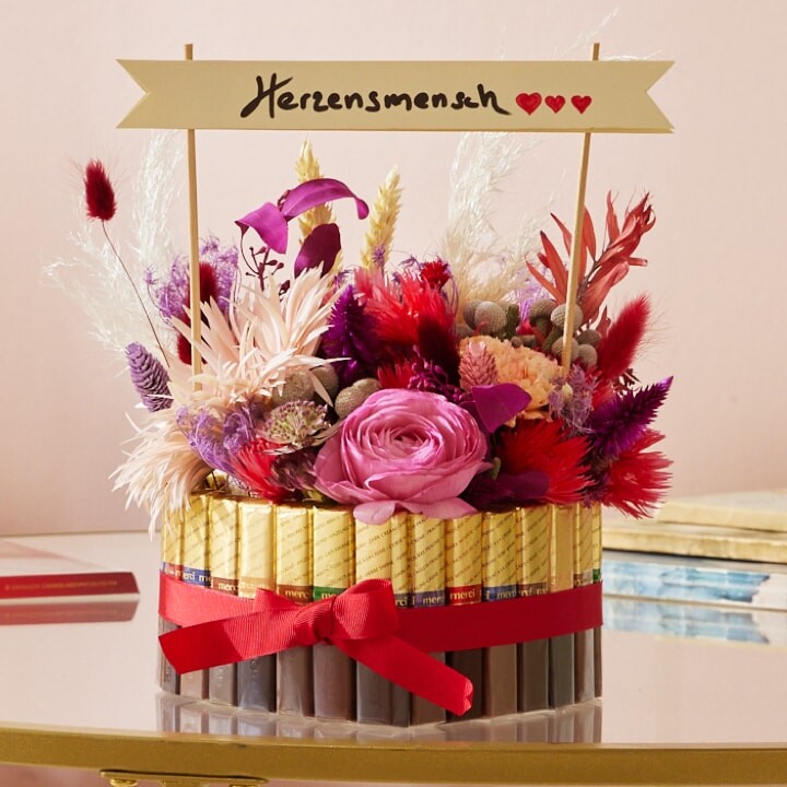 Eine Schachtel in Herzform, außen beklebt mit merci Finest Selection, innen Blumen sowie ein Papierfähnchen mit der Aufschrift: Herzensmensch